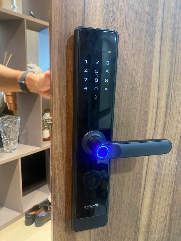 Mặt ngoài khóa vân tay Avolock AV-64 bản đen lắp đặt thực tế trên cửa gỗ căn hộ: bàn phím cảm ứng phát sáng, cảm biến vân tay trên tay nắm